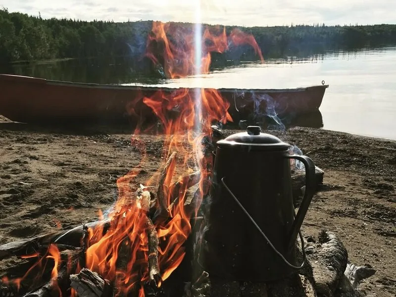 An enamel kettle beside a driftwood beach fire, a red canoe and calm lake behind
