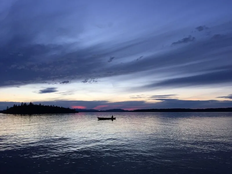 A lone canoe silhouetted on glassy water at blue hour, the last pink light behind a treed island