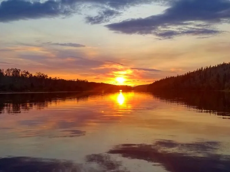 A blazing orange sunset mirrored on a calm northern lake between spruce-lined shores