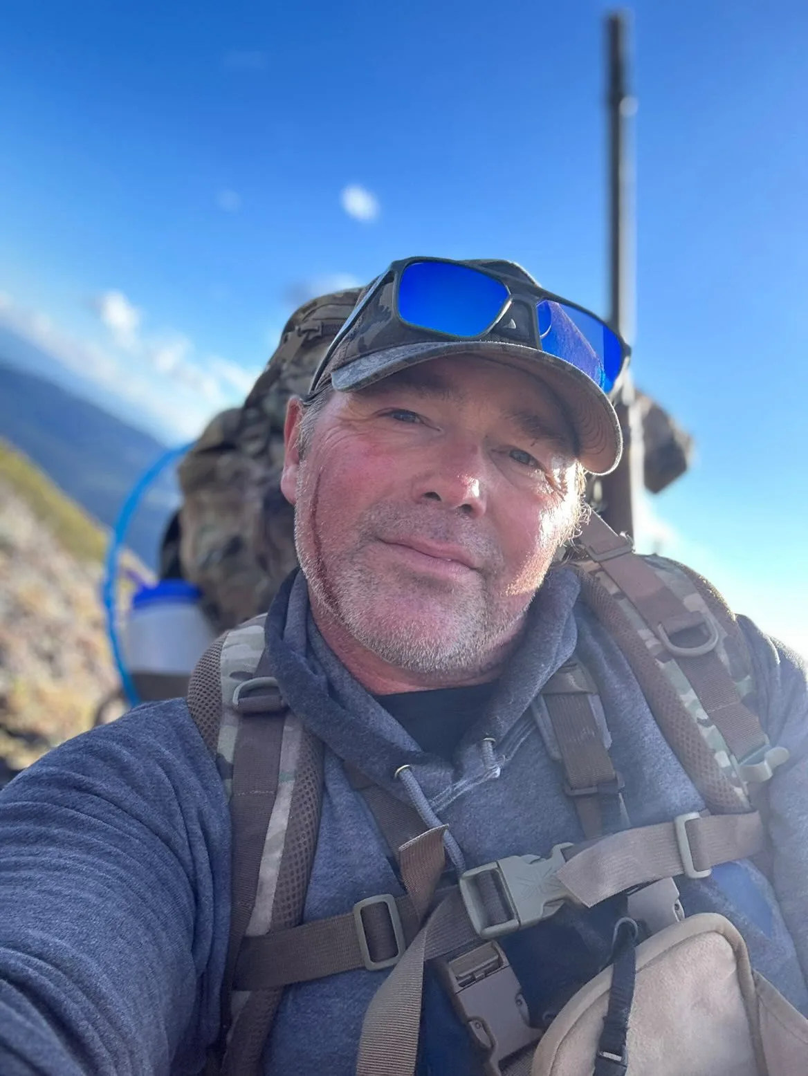 Mike Hawkridge on a mountain ridge, ballcap and pack straps, alpine ridgeline behind