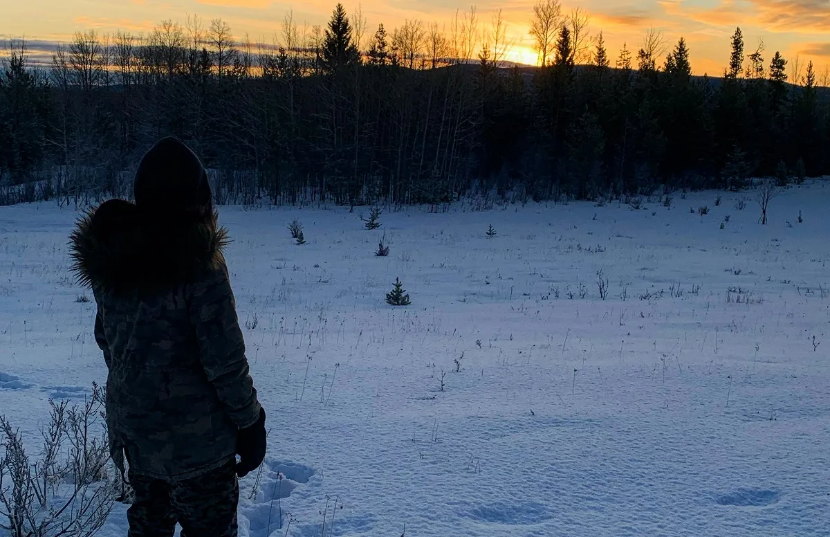 A figure in a fur-trimmed parka hood crossing untouched snow toward an amber sunset over the treeline